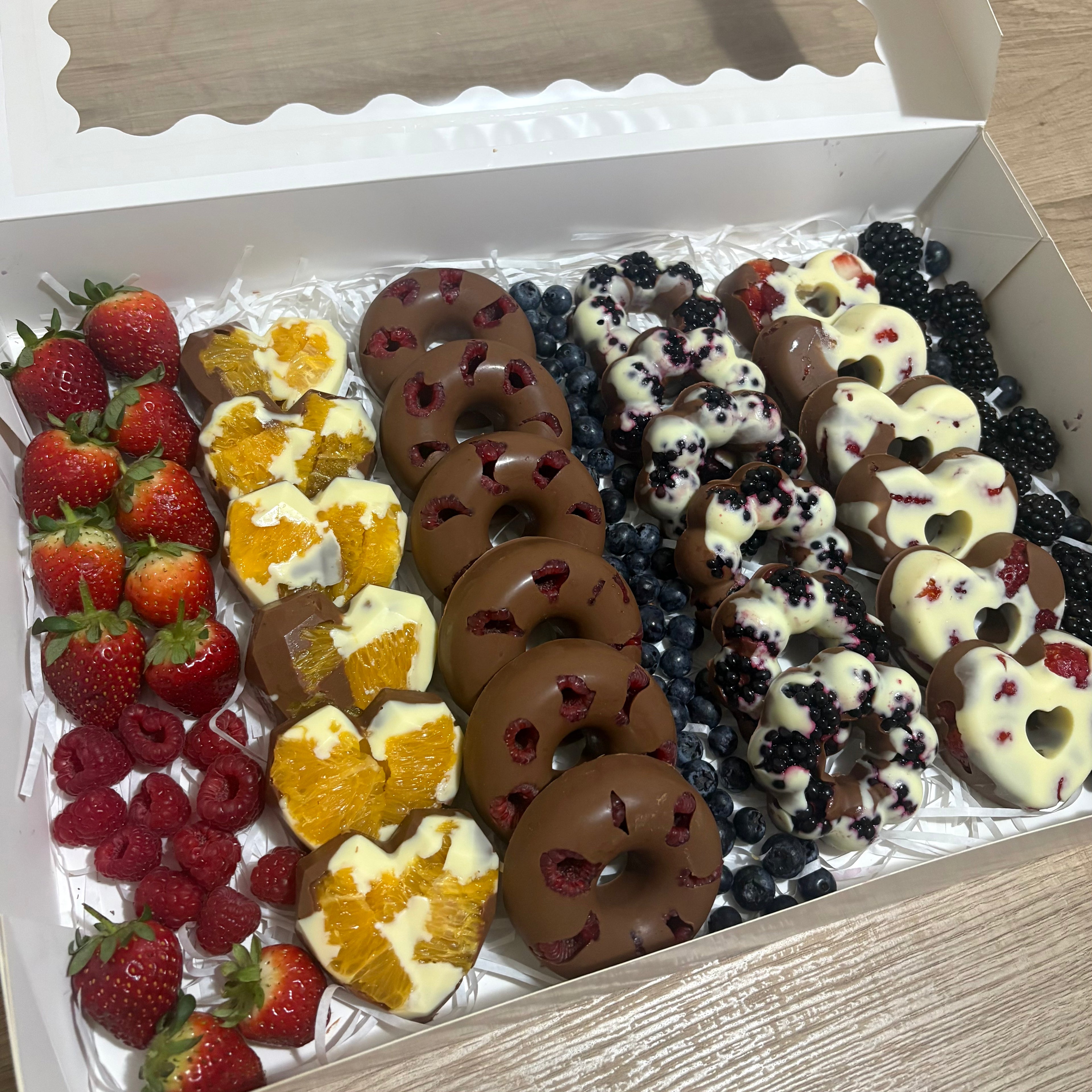 A gift box of assorted gourmet treats, including fresh strawberries, raspberries, blueberries, blackberries, chocolate-covered donuts with berry pieces, orange halves dipped in white chocolate, and heart-shaped white chocolate cookies with berry and fruit toppings.