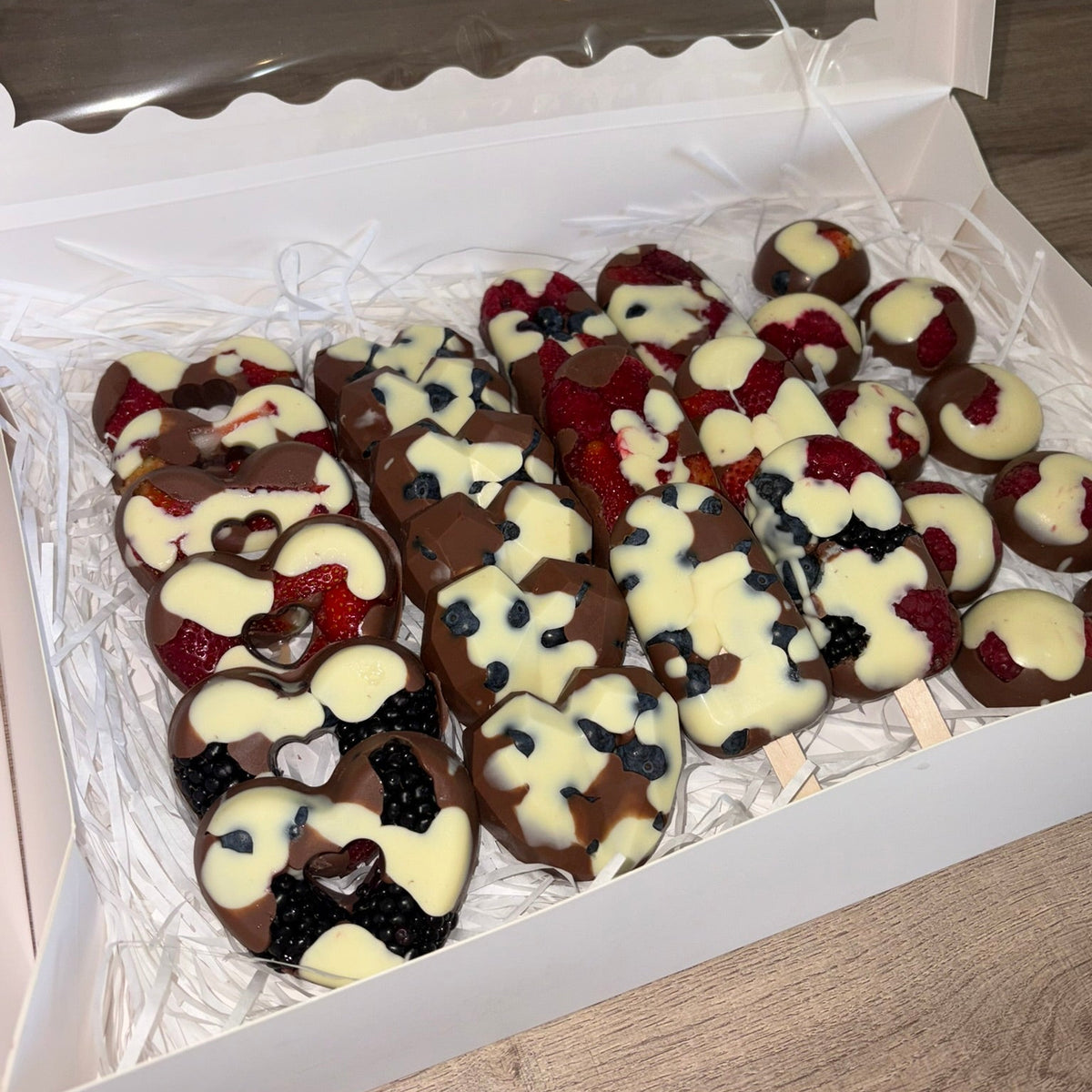 A box of assorted chocolate-covered fruit skewers, including strawberries, blueberries, and blackberries, decorated with white chocolate drizzle and heart-shaped chocolate details, presented in a white gift box.