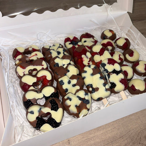 A box of assorted chocolate-covered fruit skewers, including strawberries, blueberries, and blackberries, decorated with white chocolate drizzle and heart-shaped chocolate details, presented in a white gift box.